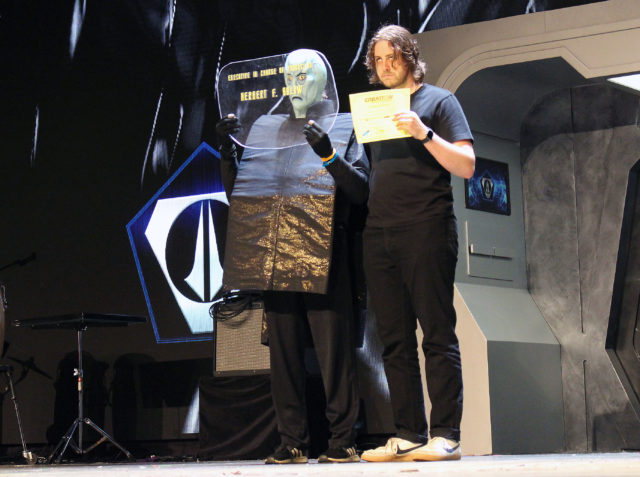 The executive in charge of production Herber F. Solow's title card gets his winning certificate from judge Noah Averbach-Katz, who is trying to do his best impression of the Balok puppet